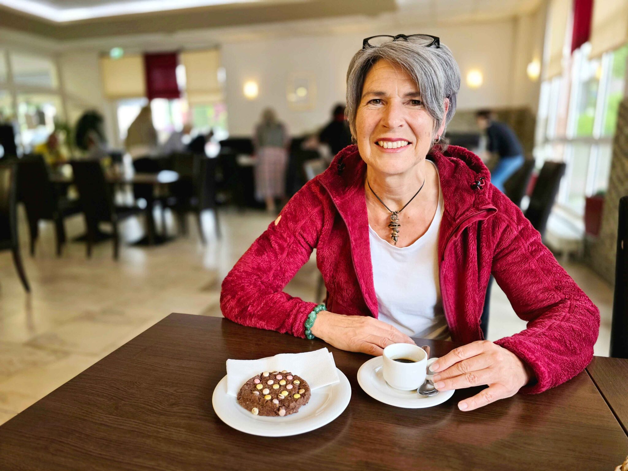 Eine lächelnde Frau mit grauen Haaren und roter Jacke sitzt im Café vor einem Espresso und Keks.