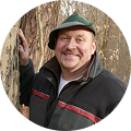 Porträt von Dirk Strauch-Förster in Arbeitskleidung und grünem Hut vor einem Baumstamm.