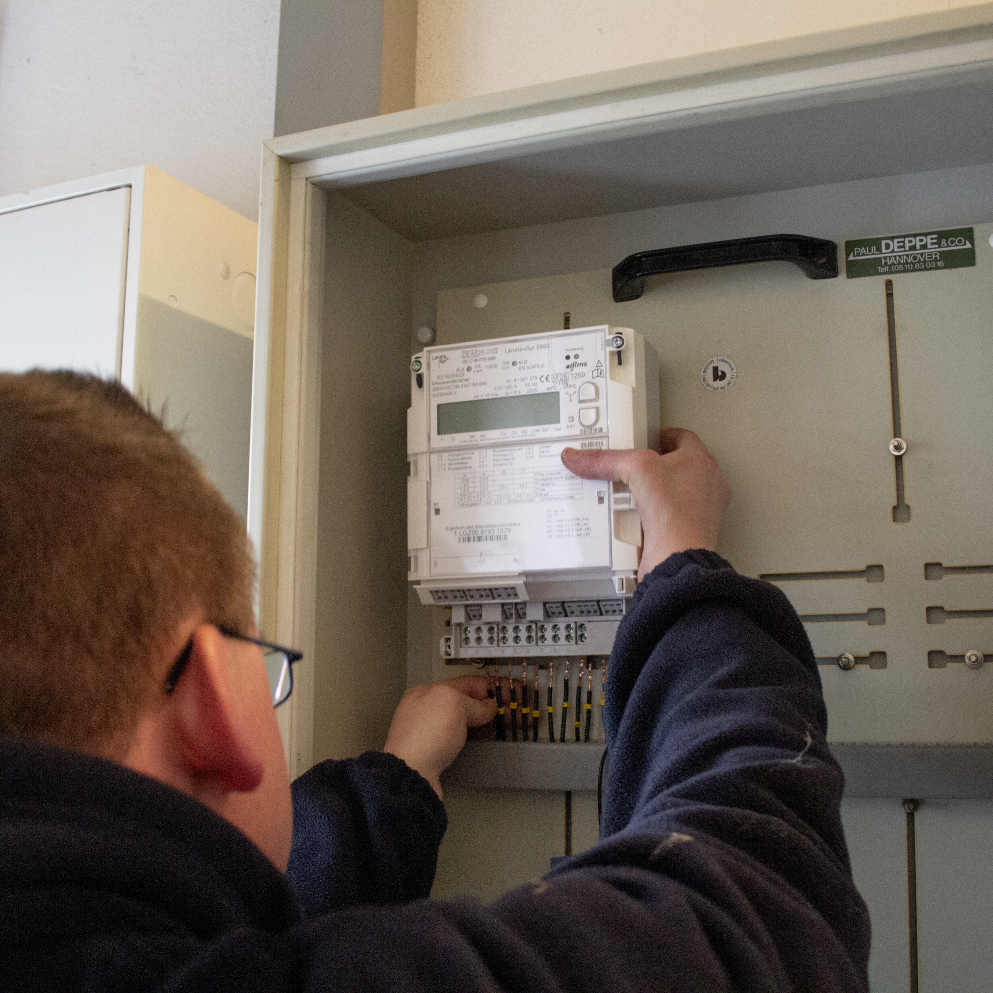 Person mit Brille und dunklem Pullover arbeitet an einem geöffneten Stromzähler in einem grauen Schaltkasten