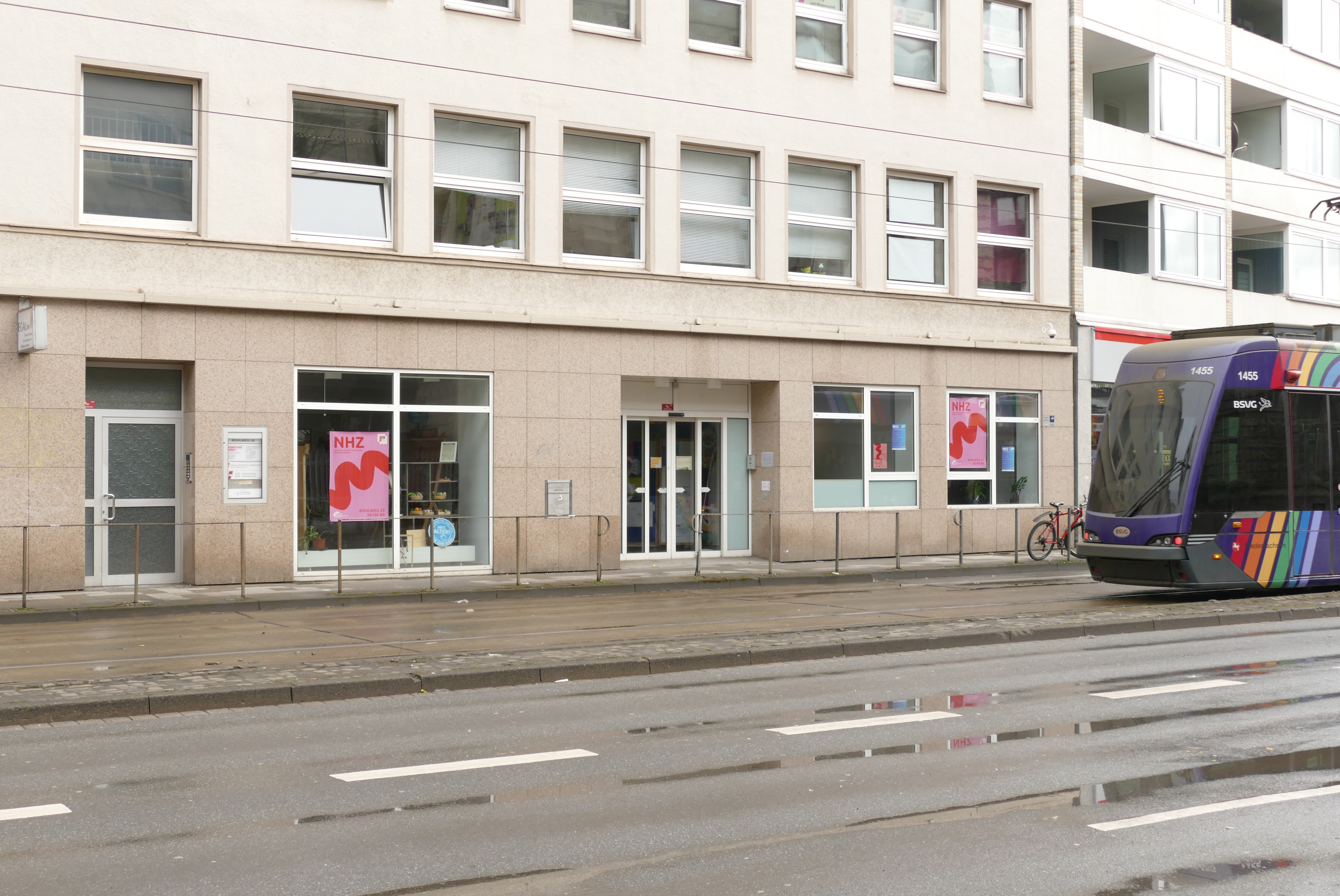 Straßenansicht einer Gebäudefront mit rosa „NHZ“-Plakaten. Rechts eine bunte Straßenbahn auf nasser Straße.